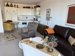 A living room with a sofa, armchair, dining table, and kitchen area in the background.