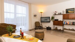 Uma sala de estar com sofá, uma mesa de jantar com vela, televisão, estante de livros e janela com cortinas.