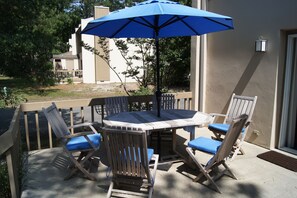 An outdoor patio with a round table and four wooden chairs with blue cushions, a blue umbrella, and a view of a building and trees in the background. - Enjoy this comfy 3 bedroom home & walk, bike or take the trolley to the beach! (Bethany Beach)