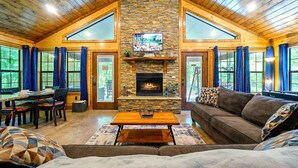 A cozy living room with a stone fireplace, wooden ceiling, and large windows. - The Blue Heron, HIDDEN IN THE WOODS YET CLOSE TO TOWN (Broken Bow)