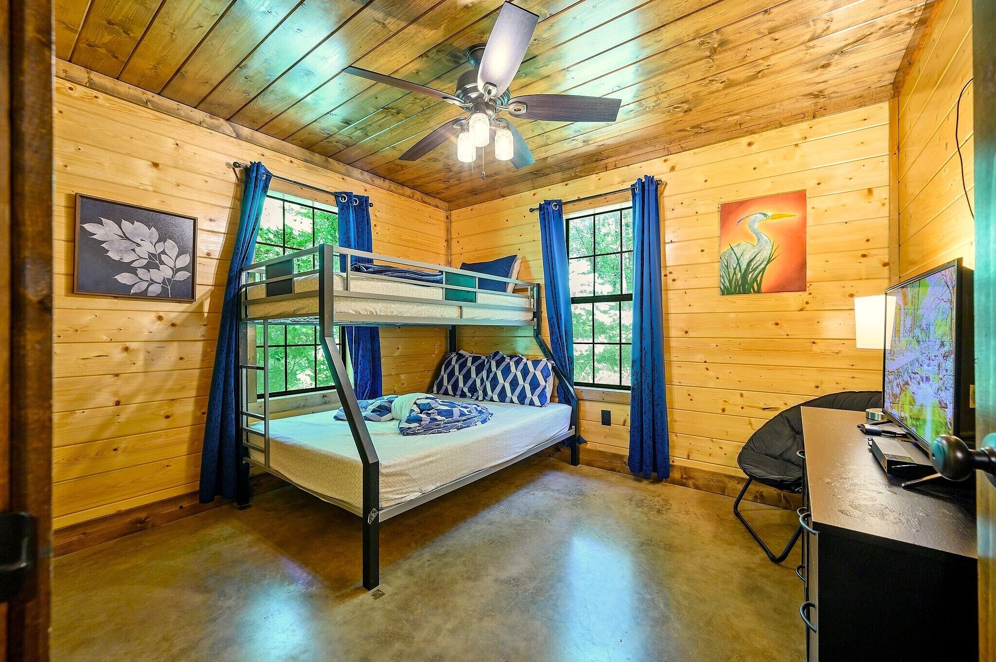 Une chambre dans un chalet en bois, avec un lit superposé, un ventilateur de plafond, un bureau avec un ordinateur et une fenêtre avec des rideaux.