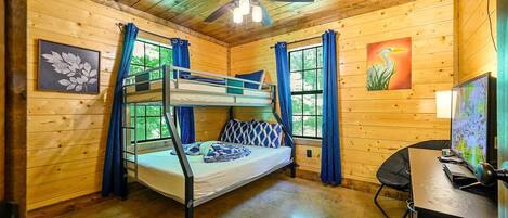 Une chambre dans un chalet en bois, avec un lit superposé, un ventilateur de plafond, un bureau avec un ordinateur et une fenêtre avec des rideaux.