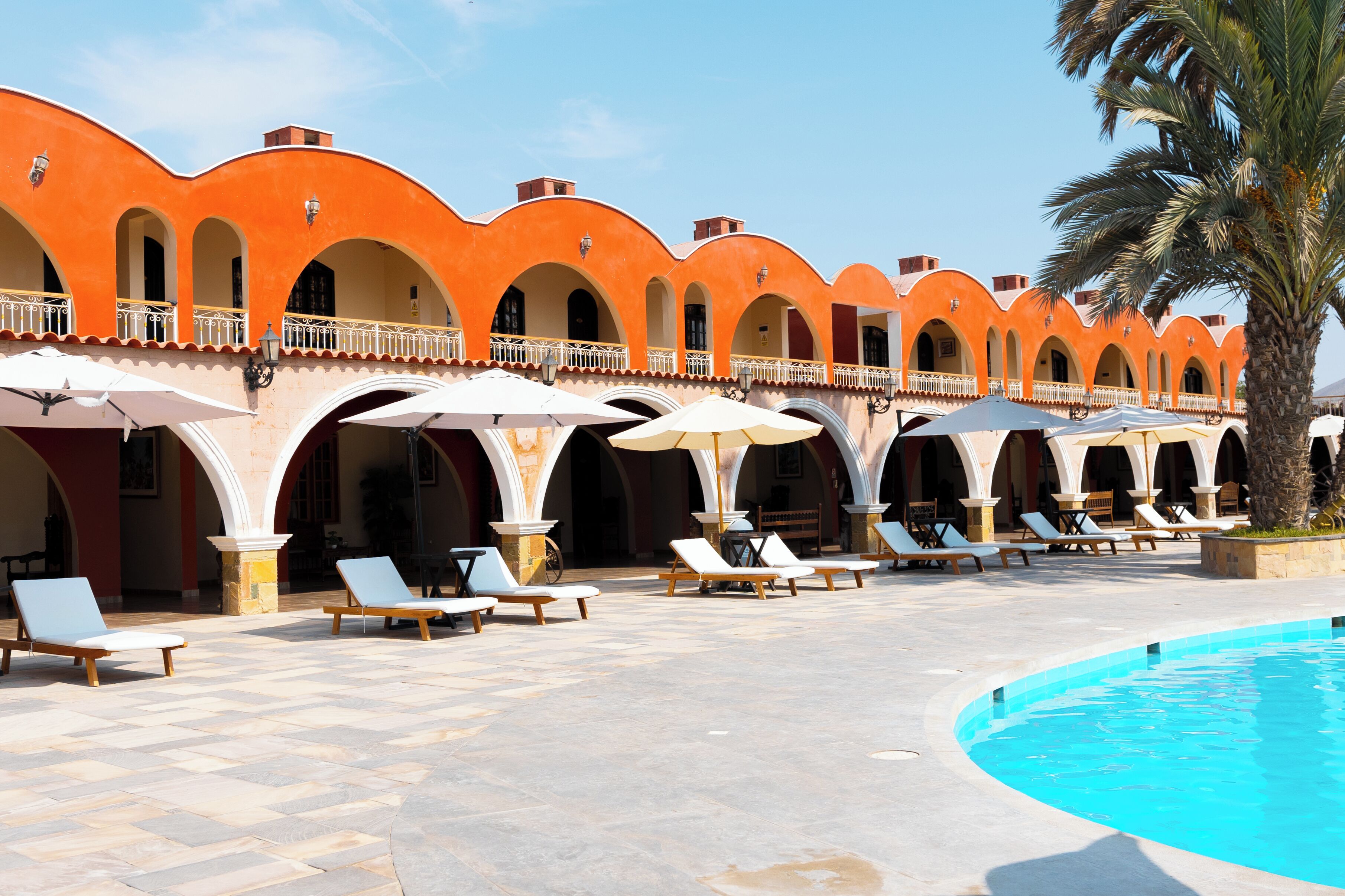 Un área junto a la piscina con tumbonas y sombrillas, una palmera y un edificio con arcos y suelo de baldosas.