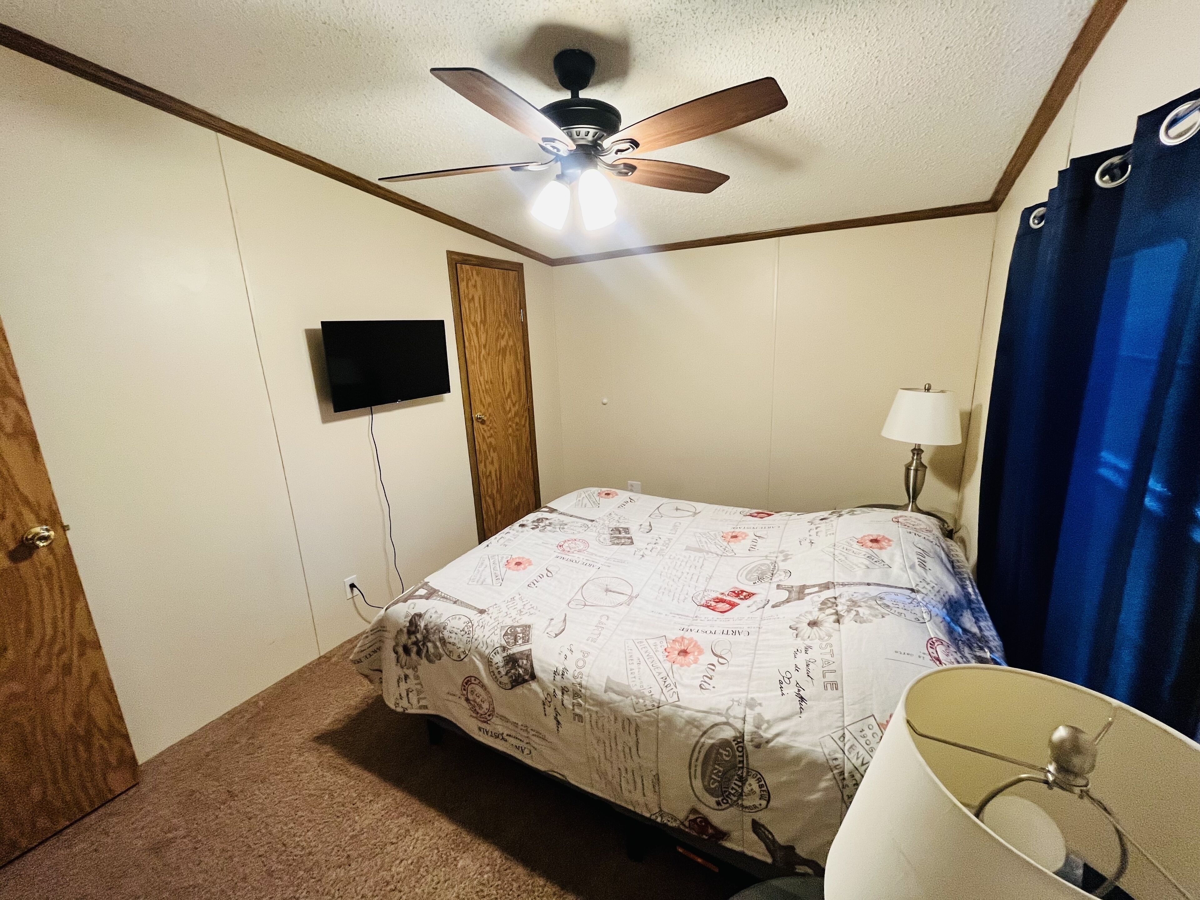 Un dormitorio con cama, ventilador de techo, televisión montada en la pared, lámpara y ventana con cortinas azules.