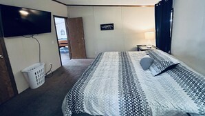Hotellihuone, jossa on sänky, televisio, seinään kiinnitetty valaisin, ovi ja pyykkikori.
