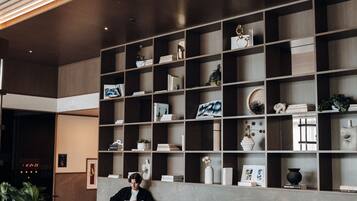 A modern lobby with a central seating area, a wall of shelves with various items, and a ceiling with linear lighting.