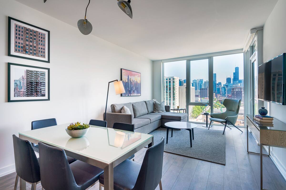 A modern living room with a glass dining table, gray sofa, and a view of the city skyline.