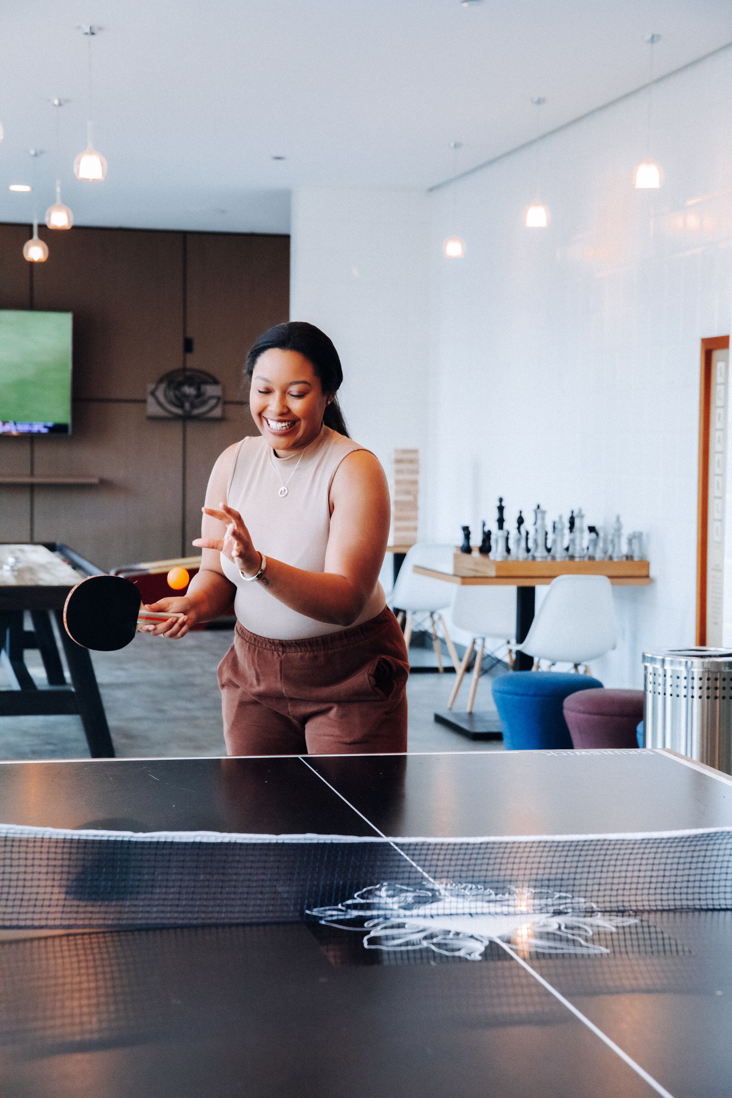 A person playing table tennis in a modern room with a ping pong table, a chessboard, and a television in the background.