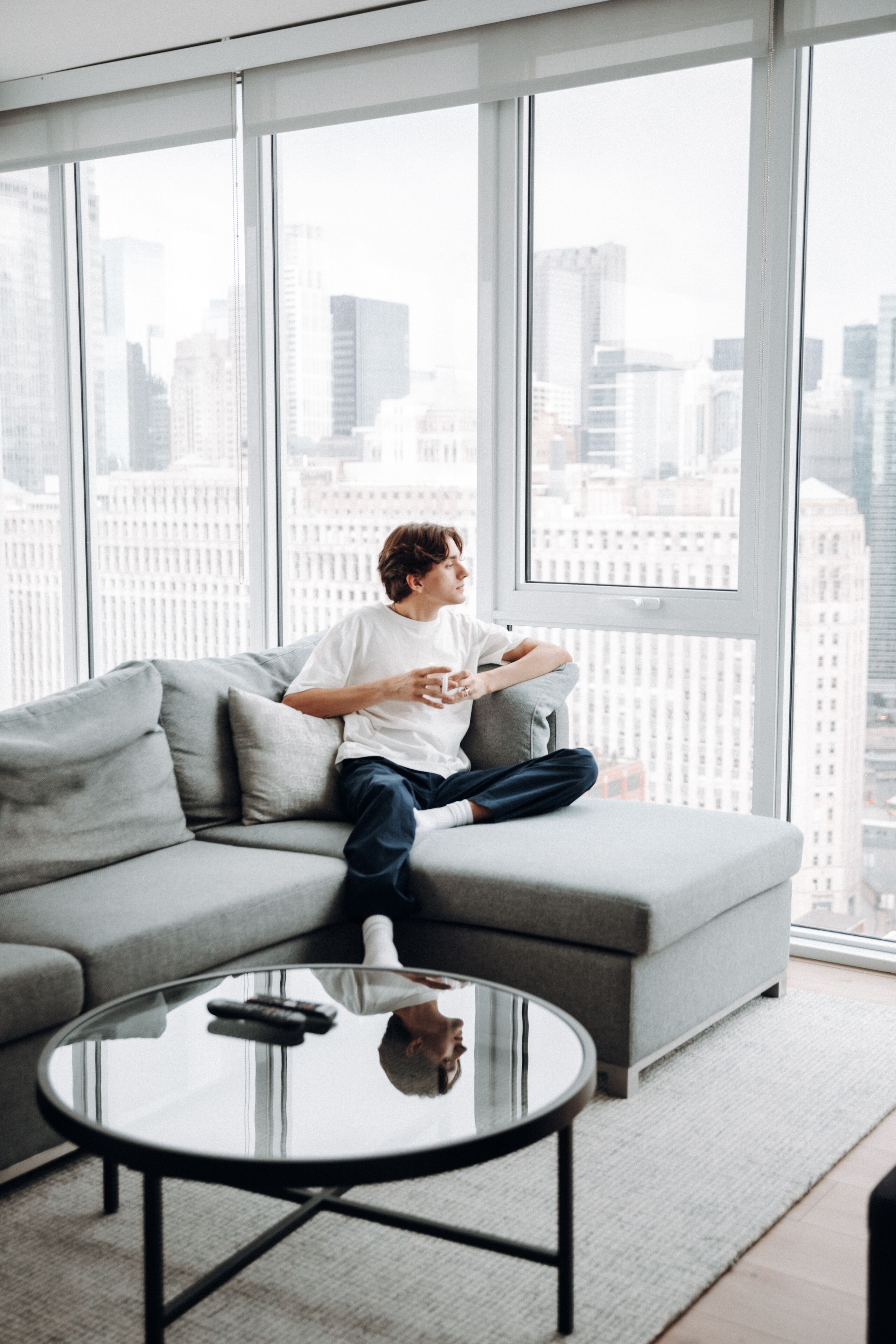 Una persona sentada en un sofá gris en una habitación moderna con un ventanal grande, una mesa de centro redonda de cristal y un control remoto sobre la mesa.