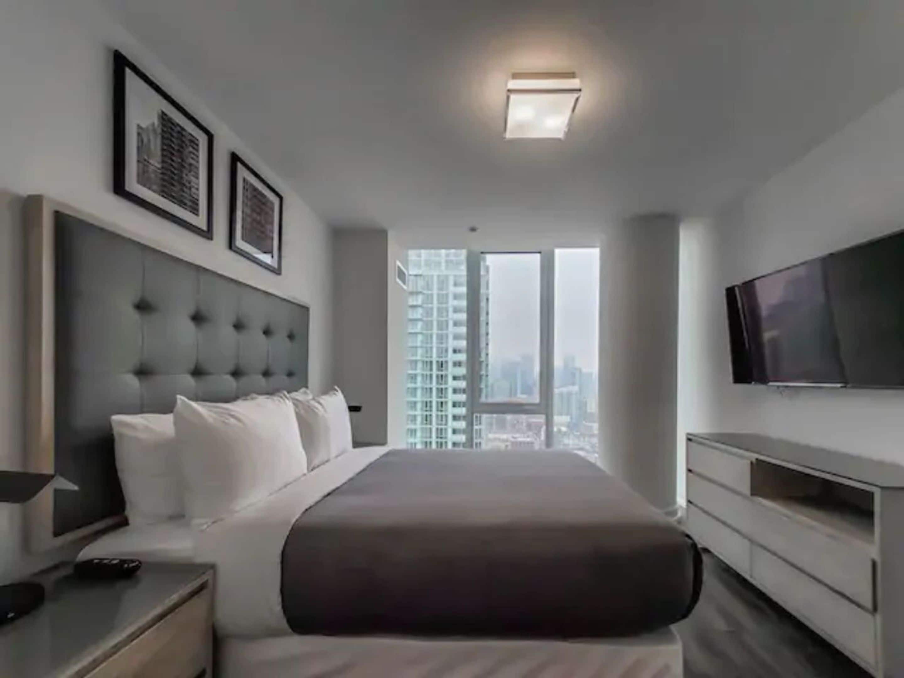 Quarto de hotel moderno com uma cama grande, televisão de tela plana, mesa de cabeceira e vista para a cidade pela janela.