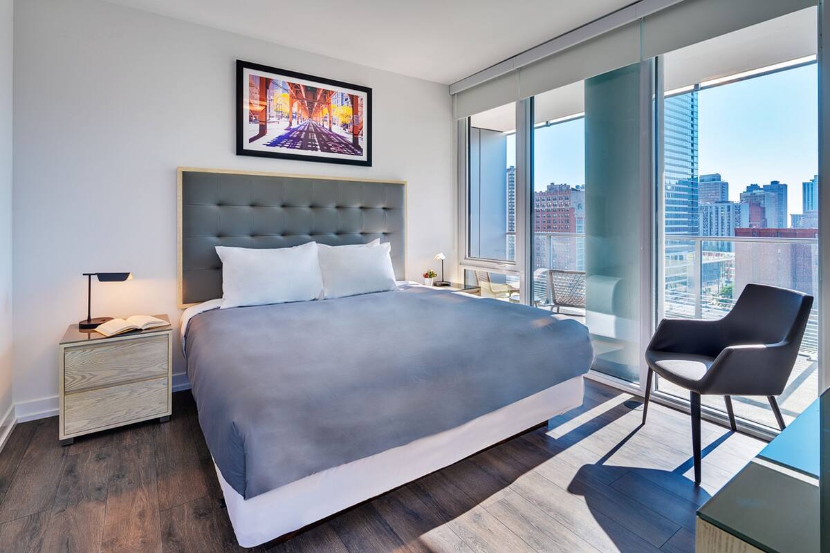 A modern hotel room with a large bed, a nightstand, a chair, and a city view through large windows.