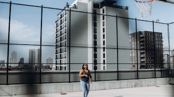 Un terrain de basket en plein air, entouré d’une clôture en treillis métallique, avec un panier de basket et une vue sur la ville en arrière-plan.