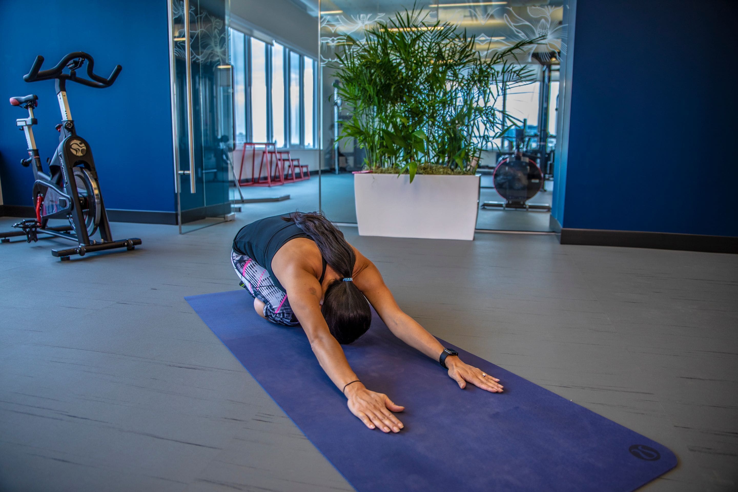 Une personne effectue une posture de yoga sur un tapis dans une salle de sport, avec des appareils d’exercice et une grande plante en arrière-plan.