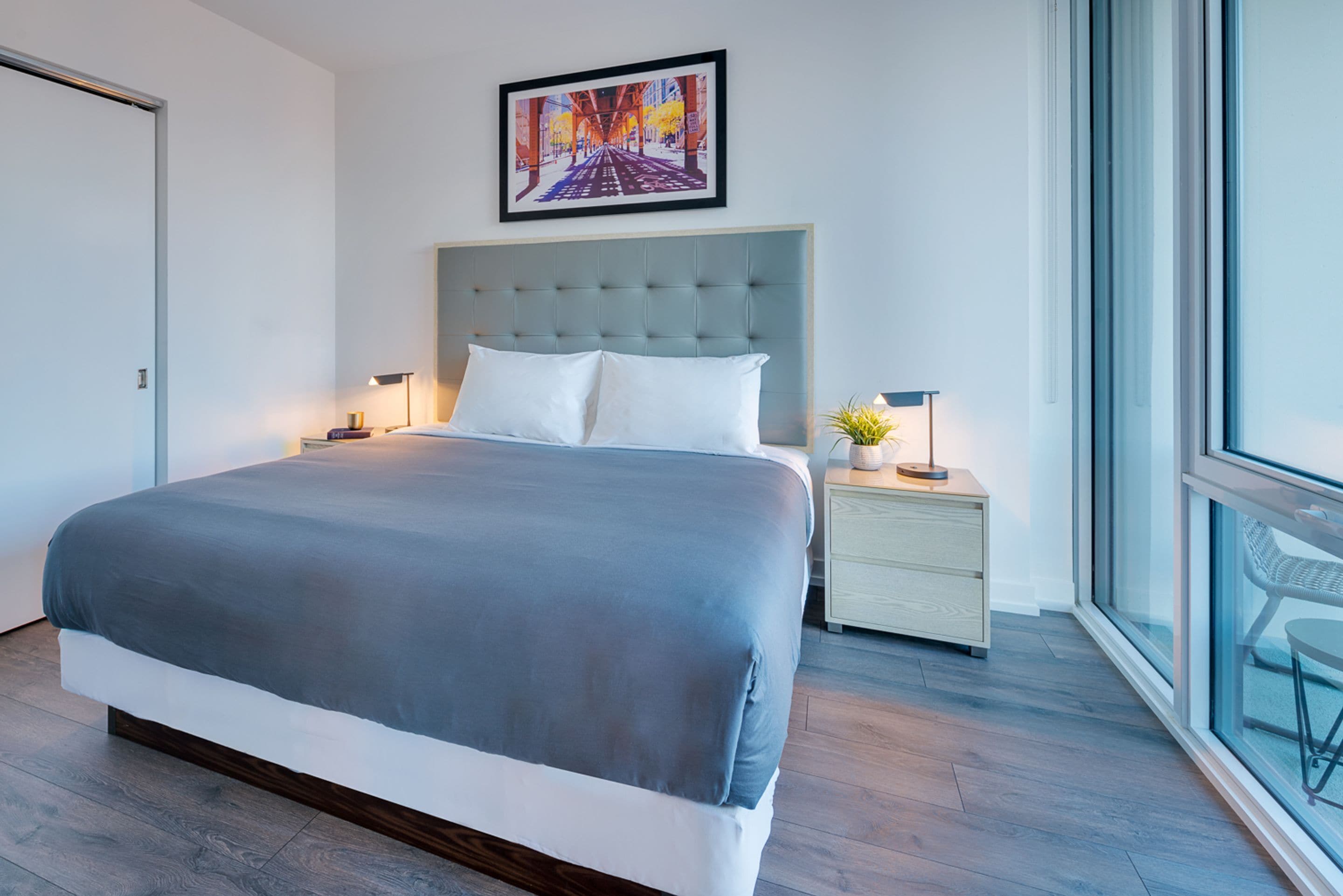 A modern hotel room with a large bed, a headboard, a bedside table with a lamp, and a window offering a view.