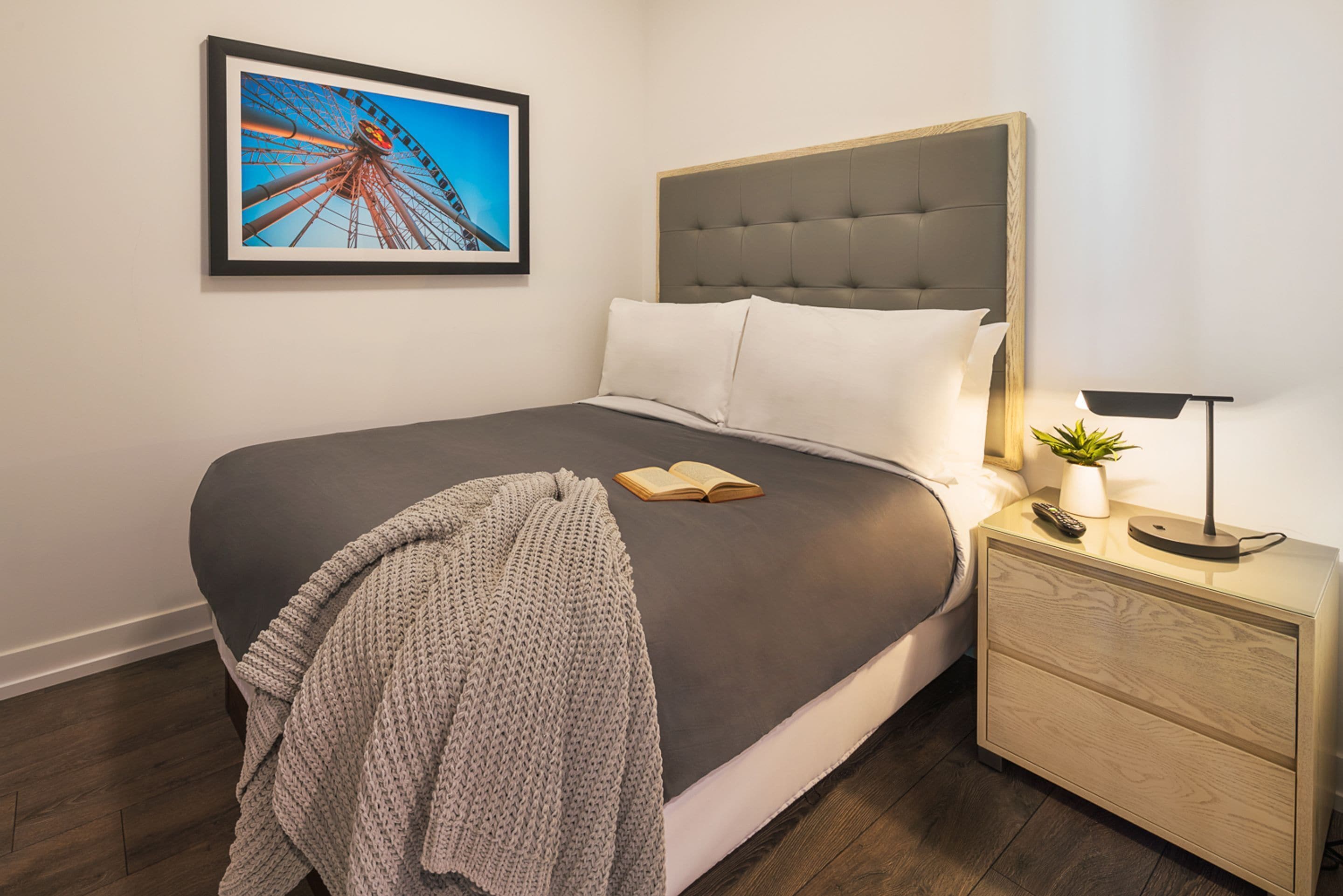 A neatly made bed with a grey blanket, a framed picture of a Ferris wheel on the wall, and a bedside table with a lamp and a plant.