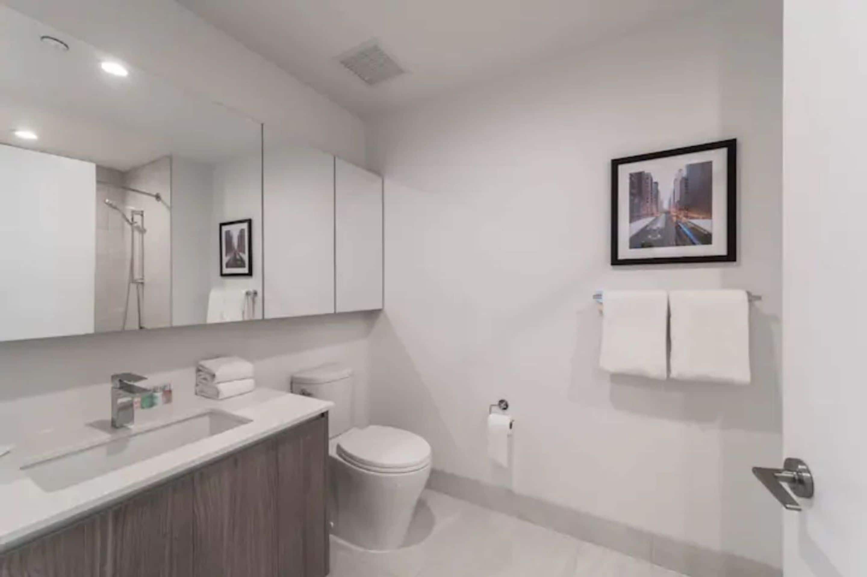 Ein modernes Badezimmer mit Waschbecken, Spiegel und Toilette in Weiß. An der Wand hängt ein Bild im Rahmen und ein Handtuch an einem Haken.