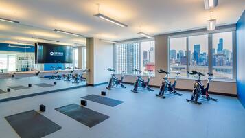 A modern fitness center with stationary bikes, a large mirror, and a city view through large windows.