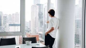 A person standing by a window in a modern office with a city view, a table with a bowl and a spoon, and a chair.