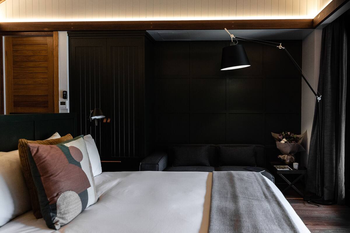 A modern bedroom with a neatly made bed, a bedside table with a lamp, and a dark wall with a wooden panel.
