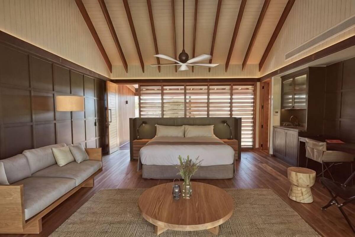 A bedroom with a bed, a sofa, a coffee table, a ceiling fan, and a view of the outside.