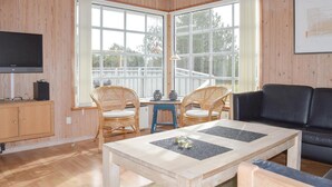 Una sala de estar acogedora con suelo de madera, un sofá de cuero negro, dos sillones de mimbre, una mesa de centro de madera y un televisor sobre un mueble de madera.