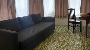 Una habitación con un sofá oscuro, una silla de madera y una mesita pequeña. Hay ventanas amplias con cortinas marrones. El suelo presenta un diseño de baldosas con motivos.