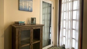 Una habitación con un armario de madera, un cuadro enmarcado en la pared, una puerta con un panel de vidrio esmerilado y una ventana con cortina blanca.