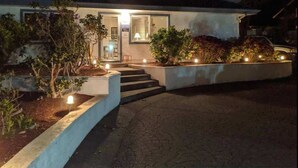 A modern house with a well-lit entrance, a staircase leading to the door, and a landscaped garden area with plants and lights. - Sparkling Clean & Private! Ocean Views! Two Full Bathrooms! [6n7] (Brookings)