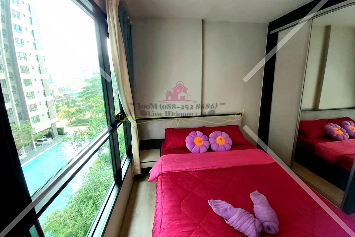 A hotel room with a large window, a bed with red bedding, two purple flower pillows, and a mirror.