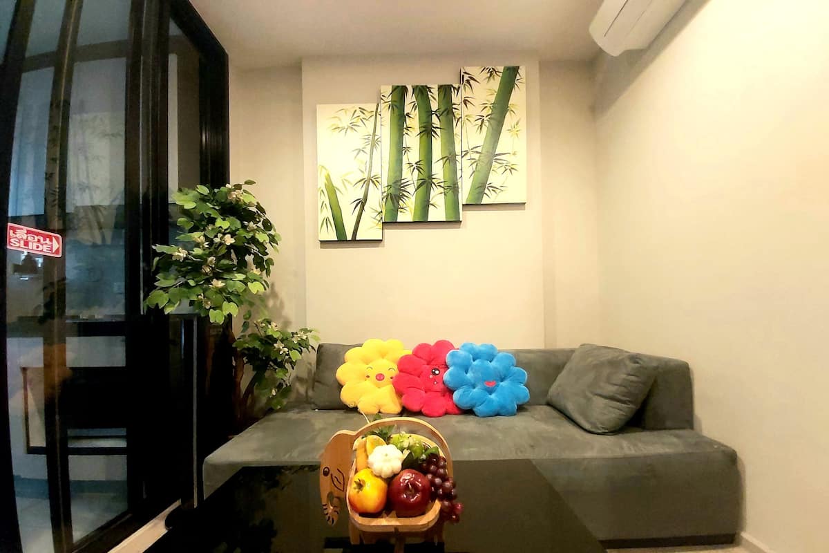 A modern living room with a grey sofa, colorful flower-shaped cushions, a wooden side table with a fruit basket, and a large window with a plant.