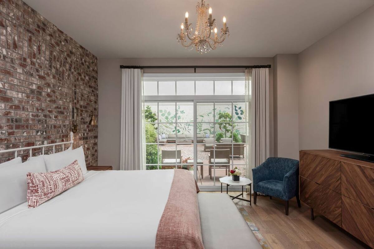 A modern hotel room with a brick wall, a large bed with white bedding, a chandelier, a TV, and a view of the outdoors.