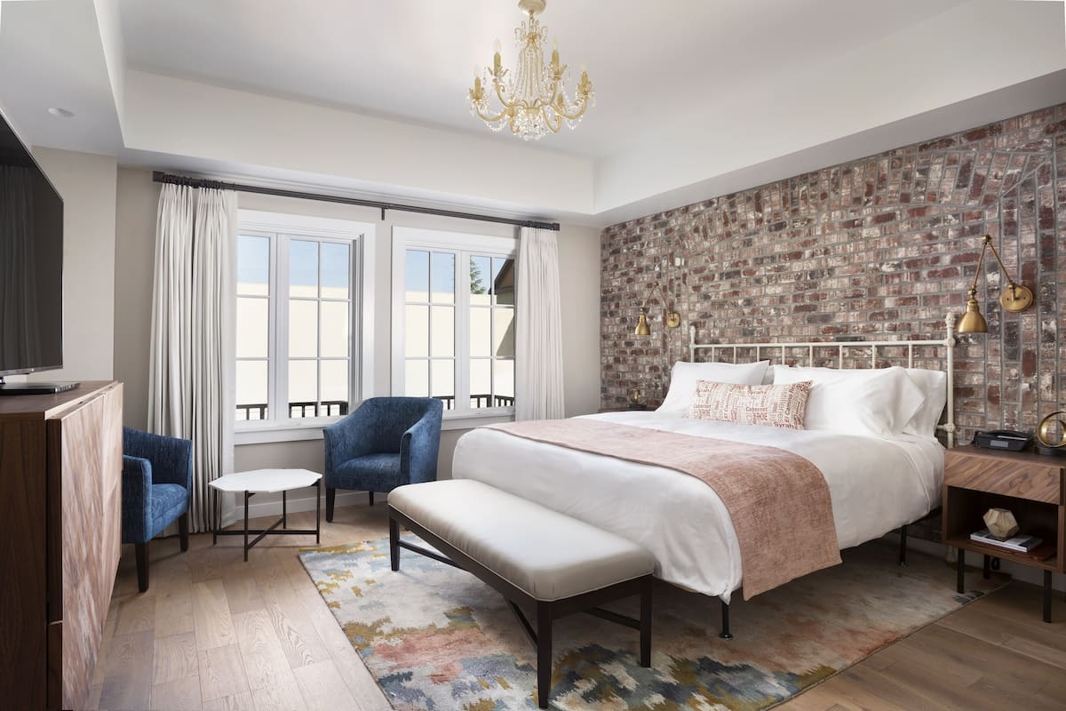 A bedroom with a large bed, a bench, a desk, a chair, a TV, and a brick-patterned wall.