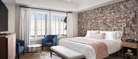 A bedroom with a large bed, a bench, a desk, a chair, a TV, and a brick-patterned wall.