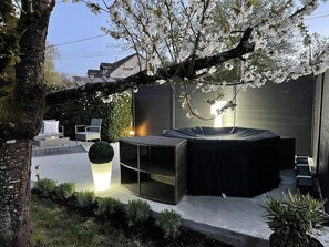 Un espacio exterior moderno con un jardín, que incluye una mesa redonda y sillas, rodeado de un árbol grande y ramas florecientes.
