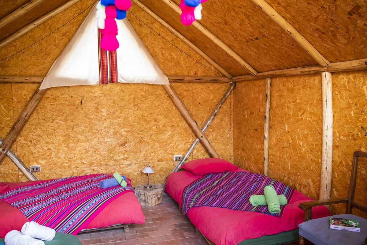 A cozy attic-style room with two beds, each with colorful bedding and pillows. There is a small wooden stool with a lamp beside one bed.