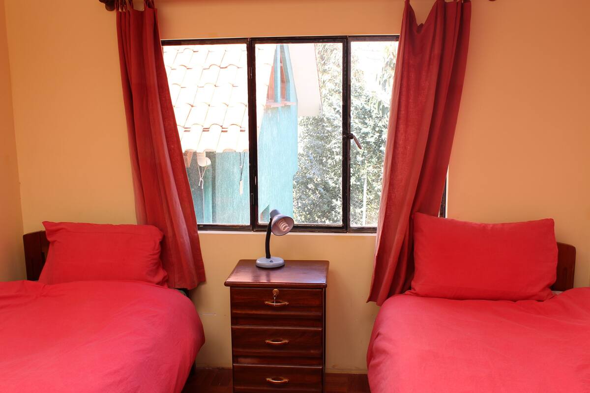 A room with two beds, a nightstand with a lamp, and a window with curtains.