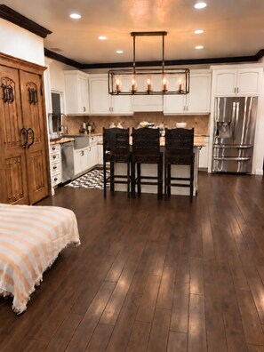 A modern kitchen with white cabinets, a central island with chairs, a large refrigerator, and a wooden wardrobe.