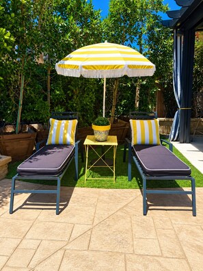 Deux fauteuils de jardin avec des coussins à rayures jaunes et blanches, une petite table sur laquelle est posé un pot jaune, et un parasol qui offre de l’ombre.
