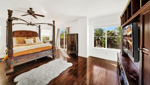 A bedroom with a four-poster bed, a ceiling fan, a large window with a view of palm trees, and a television on a wooden cabinet.