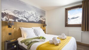 Un cuarto de hotel con una cama, una mesita de noche, una ventana con cortinas y un mural con paisaje de montaña en la pared.
