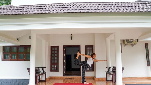 Une personne effectue une posture de yoga sur un tapis rouge, dans un espace de véranda abritée, avec des bancs en bois et un toit en tuiles.