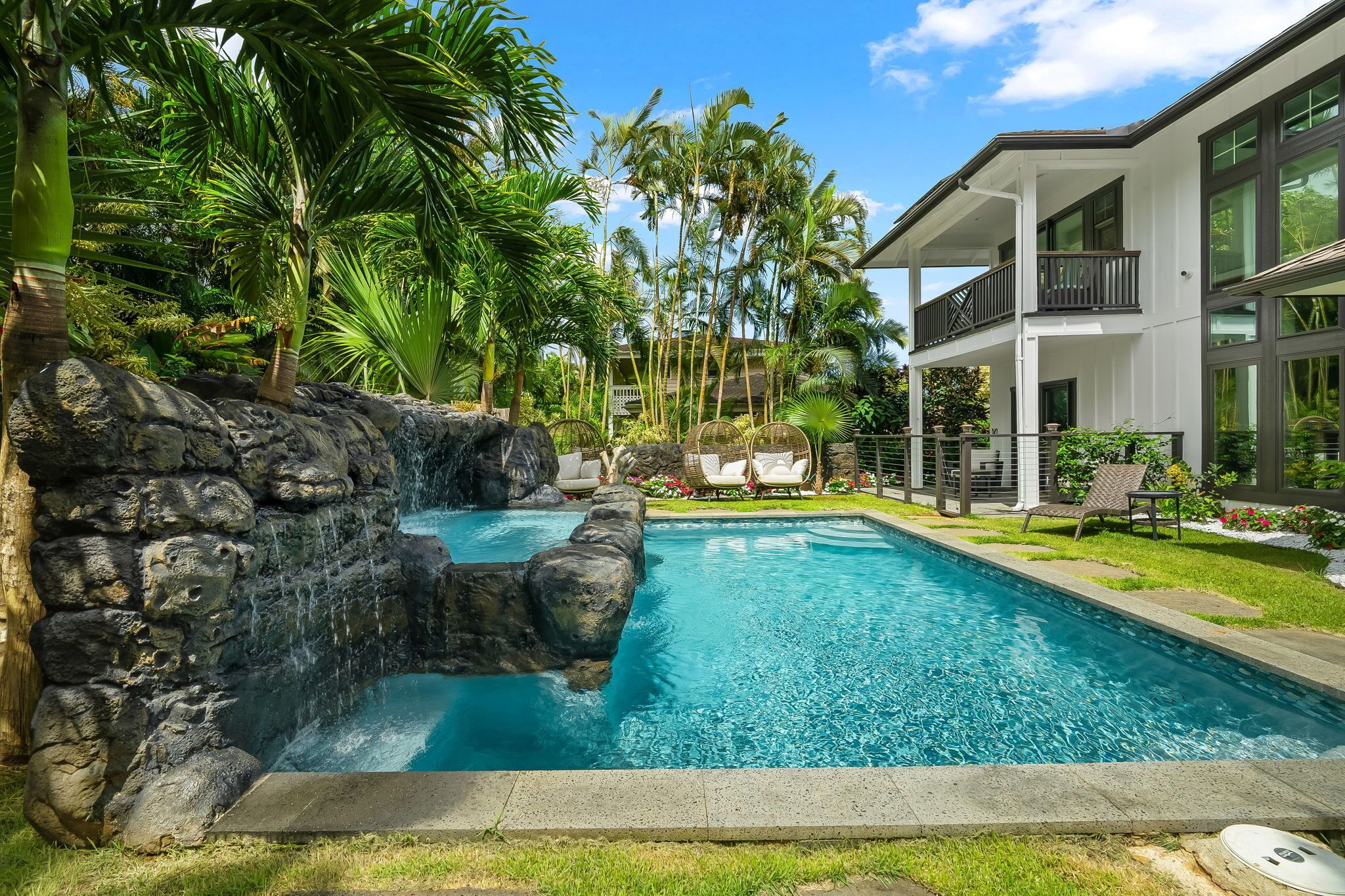 A swimming pool with a waterfall feature, surrounded by tropical vegetation and a modern building with a balcony.