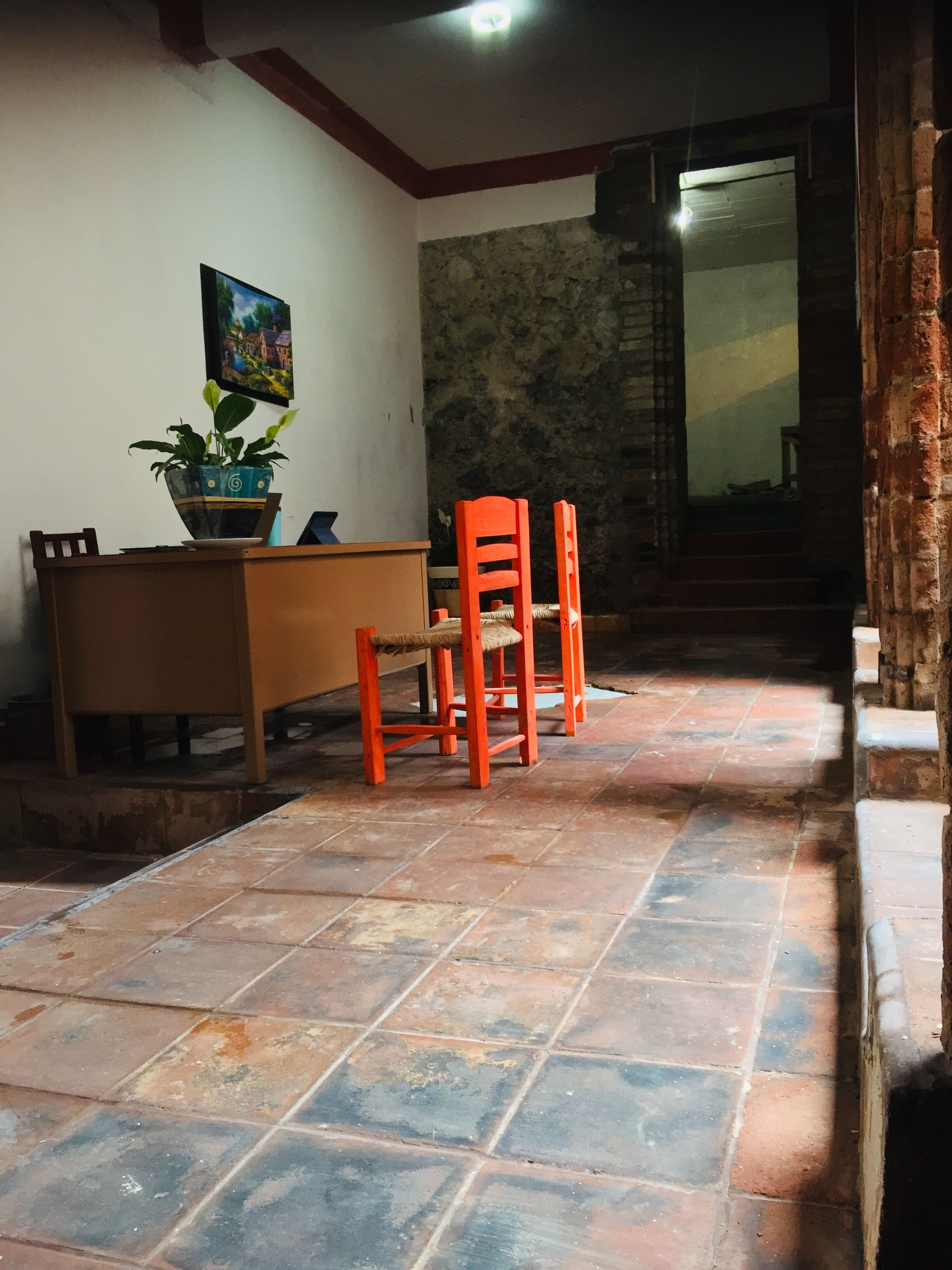 Una habitación con suelo de baldosas, un escritorio con silla, una planta en maceta y un cuadro en la pared.