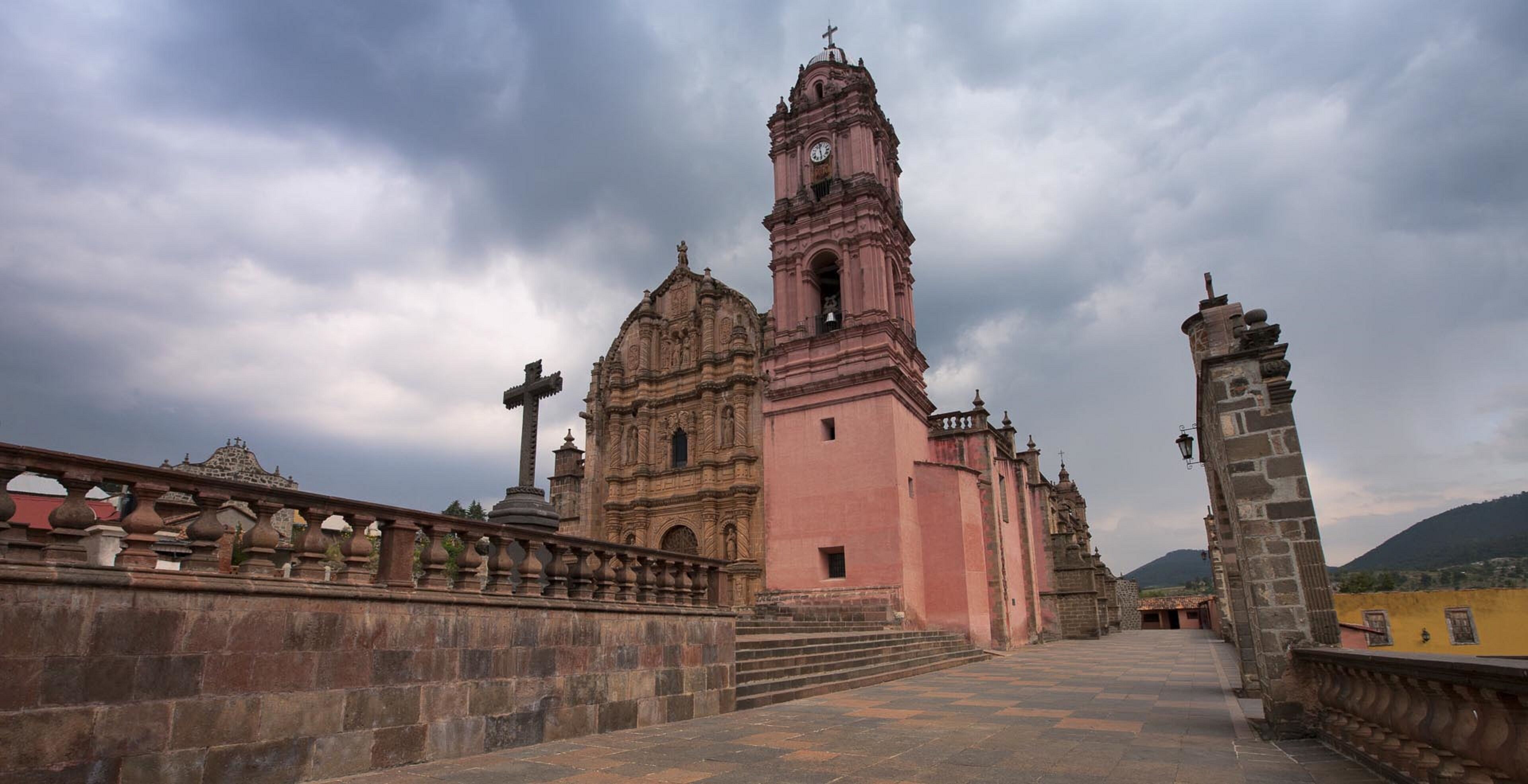 A historic church with a prominent bell tower and a large cross, featuring a grand entrance and a stone bridge in the foreground.