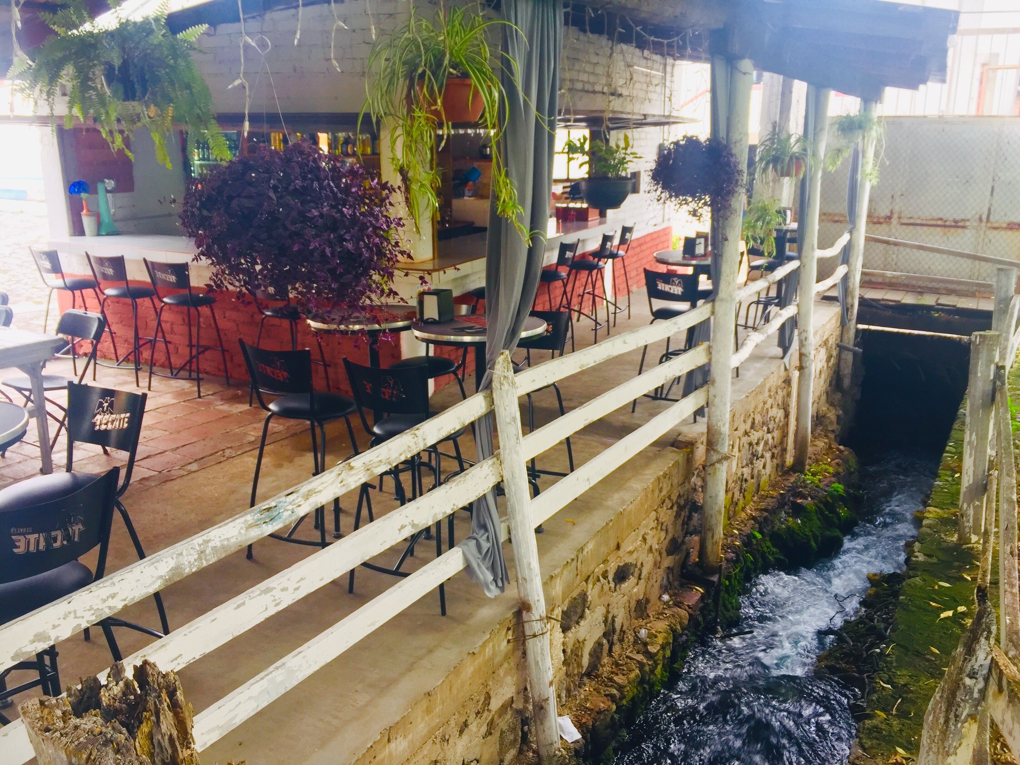 Un espacio al aire libre con mesas y sillas, barandilla blanca y un arroyo que atraviesa el lugar.