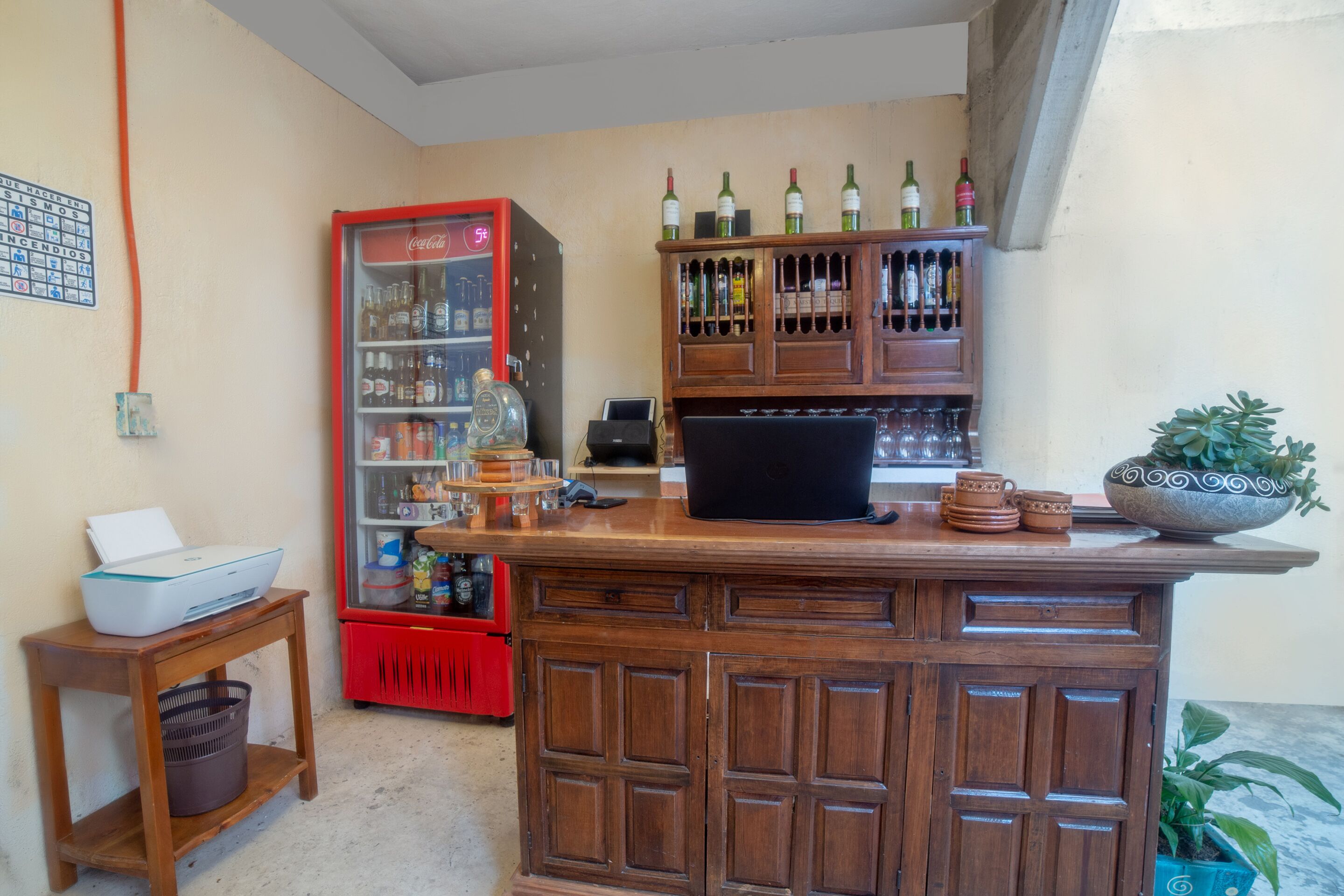 Uma área de bar com balcão de madeira, um laptop, uma geladeira com garrafas e uma pequena mesa com impressora e cesto.