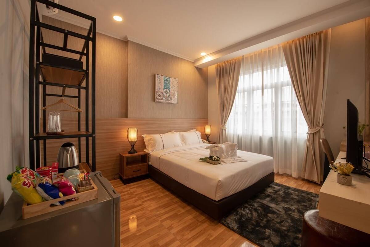 A hotel room with a bed, bedside lamps, a wooden headboard, a small table with a plant, and a window with curtains.