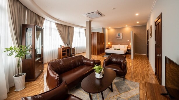 A hotel room with a brown leather sofa, a small round table with a plant, a wooden cabinet, and a bed with a white duvet. - One Vientiane Hotel & Restaurant (Vientiane)