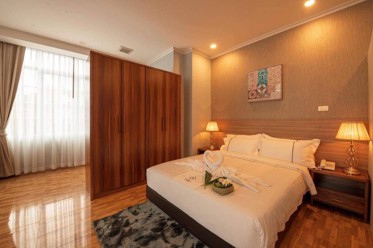 A hotel room with a wooden wardrobe, a bed with white linens, a bedside table with a lamp, and a decorative wall piece.