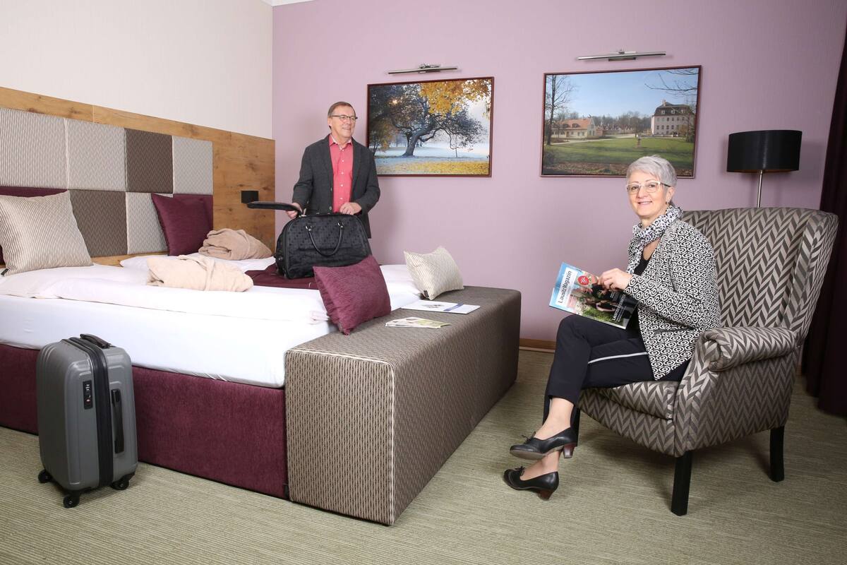 Een hotelkamer met een bed, een zithoek met een stoel, een koffer en een persoon die een tijdschrift vasthoudt.